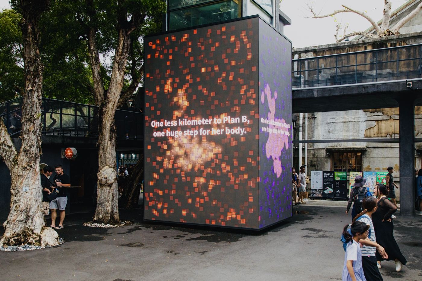 A four-sided LED cube in a Huashan courtyard showing the Taipei Plan B animation, with the line One less kilometer to Plan B, one huge step for her body