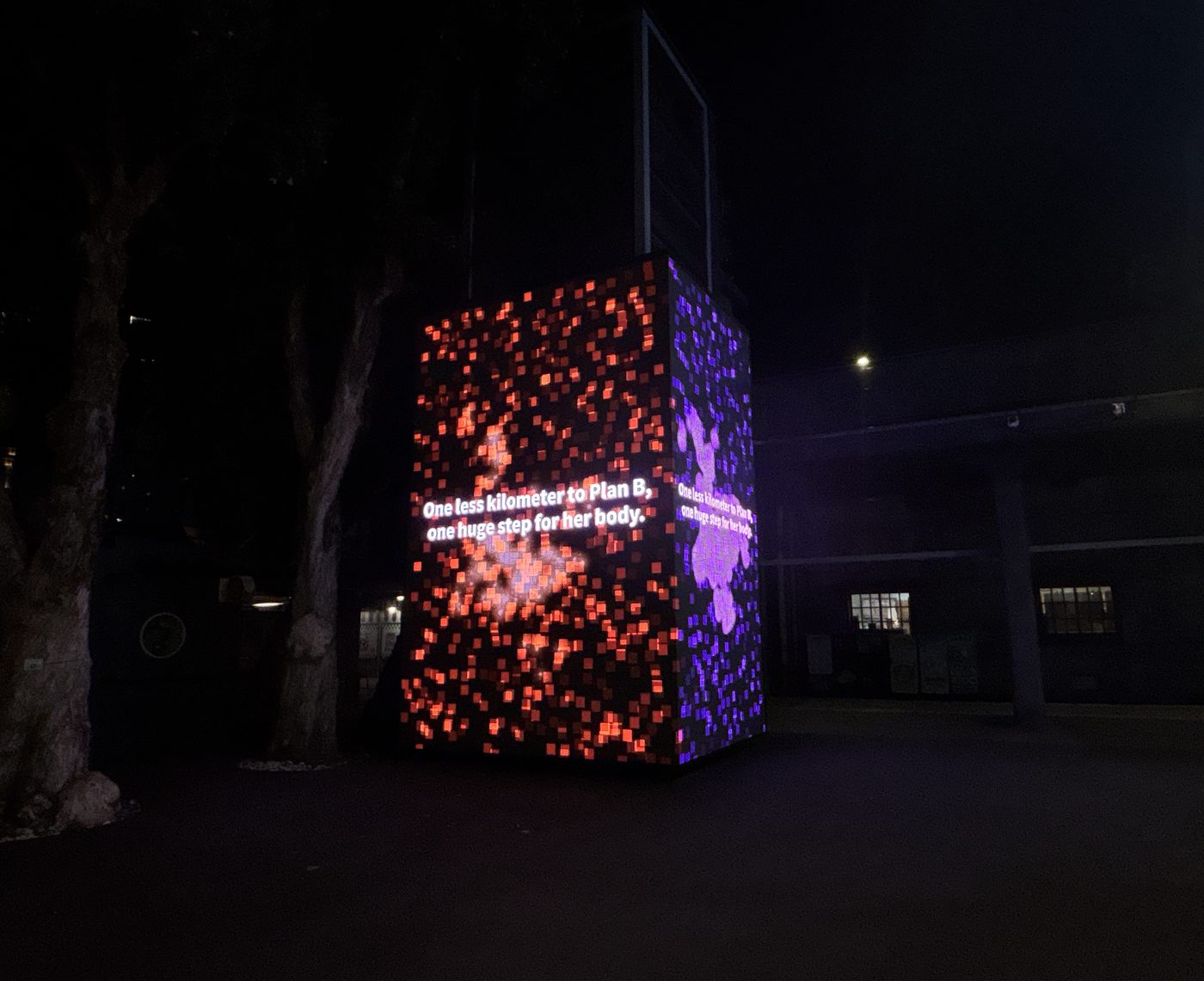 The same LED cube at night, the orange and violet pixel field glowing against a dark courtyard